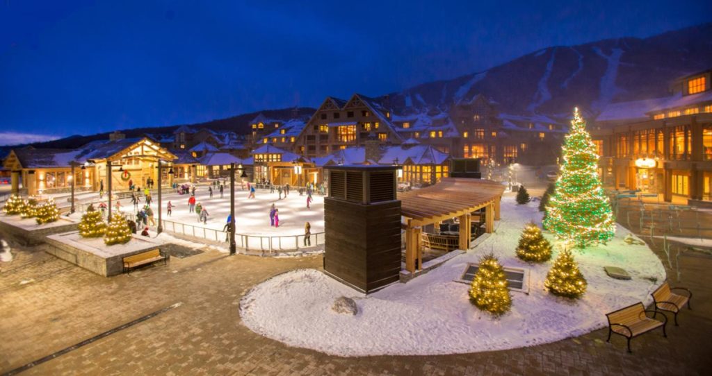 Wide angle view of holiday scene at Spruce Peak - ice skating to left and Christmas trees to right with ski slopes visible in background