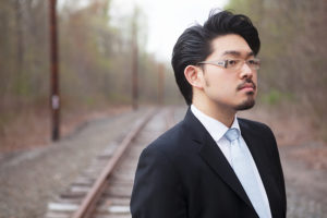 Euntaek Kim in dark suit with light blue tie and glasses standing on train tracks.
