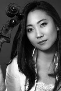 Black and white photo of Jia Kim with long brown hair and a white blouse/jacket with her cello.