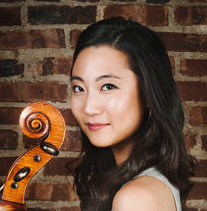 Jia Kim in light grey tank top holding her cello standing in front of a brick wall.
