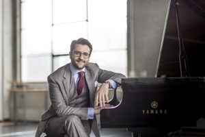 David Kaplan in a grey 3-piece siut with a blue shirt, burgundy tie, leaning on a grand piano with short brown hair mustache & beard wearing glasses, window in the background.