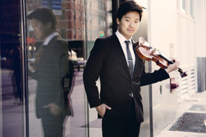 Siwoo Kim in a black suit and tie on a city street holding his violin, his reflection in the window to the left.