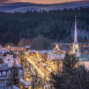 Stowe Village in Winter at twilight lit up, with mountains and sun setting in the background and Stowe Community Church steeple to the right