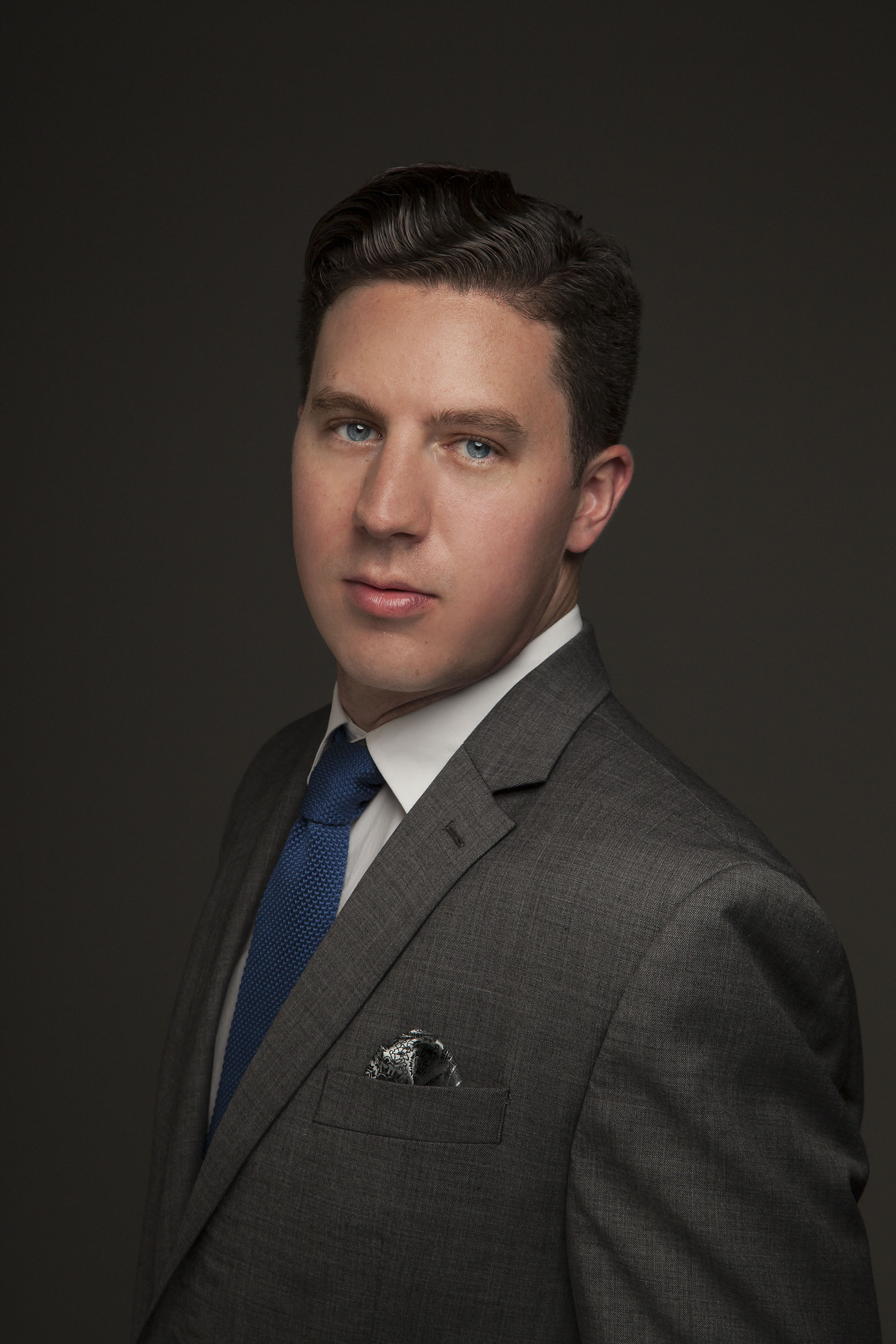 Colin Brookes in a gray suit with white shirt and blue tie against a dark gray background.
