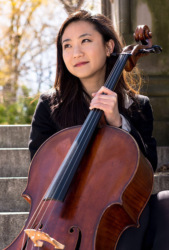 Jia Kim in a dark suit standing in front of concrete stairs with her cello, fall foliage behind her.