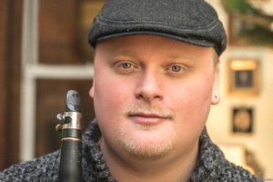 Mark Dover in gray sweater and scali cap holding his clarinet on a sidewalk.
