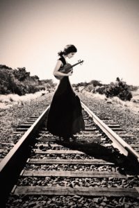 Black and white with rose tint photo - Michelle Ross in a long dark dress holding a violin on train tracks