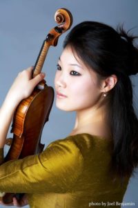 Rachel Lee Priday holding her violin in an olive silk dress and long black hair against a blue background.