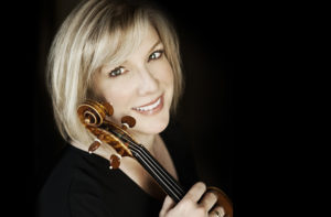 Annie Fuillard with blonde hair and black dress holding her violin with black background.