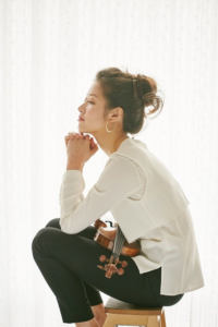 Chrystel Lee in a white top with black pants sitting on a stool with her violin in her lap against a white background.
