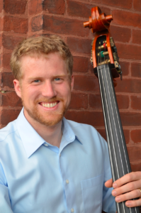 Evan Premo in a light blue collared shirt holding his bass against a brick wall.