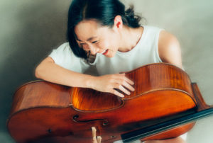 Jia Kim holding her cello on her lap, head down leaning forward laughing with a sage green mottled backgound.