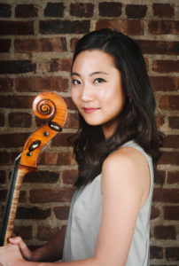 Jia Kim in light grey tank top holding her cello standing in front of a brick wall.