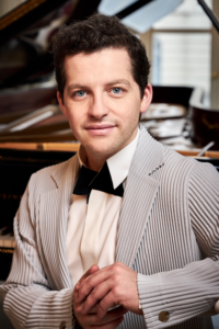 Henry Kramer sitting on a piano bench in a light gray tuxedo.