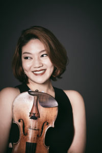 Jinjoo Cho in a blask sleeveless dress with brown bobbed hair holding her violin against a dark gray background.