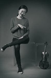 Black and white photo of Jennifer Liu sitting in a dark sweater and black pants and socks, pointing playfully at her violin propped on the floor.