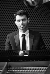 Black and white photo of Peter Dugan sitting at the piano in a dark suit with a reflection of his face in the piano top right.