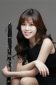 Yoonah Kim sitting holding her clarinet in a sleeveless black dress, long brown hair in front of a gray background.