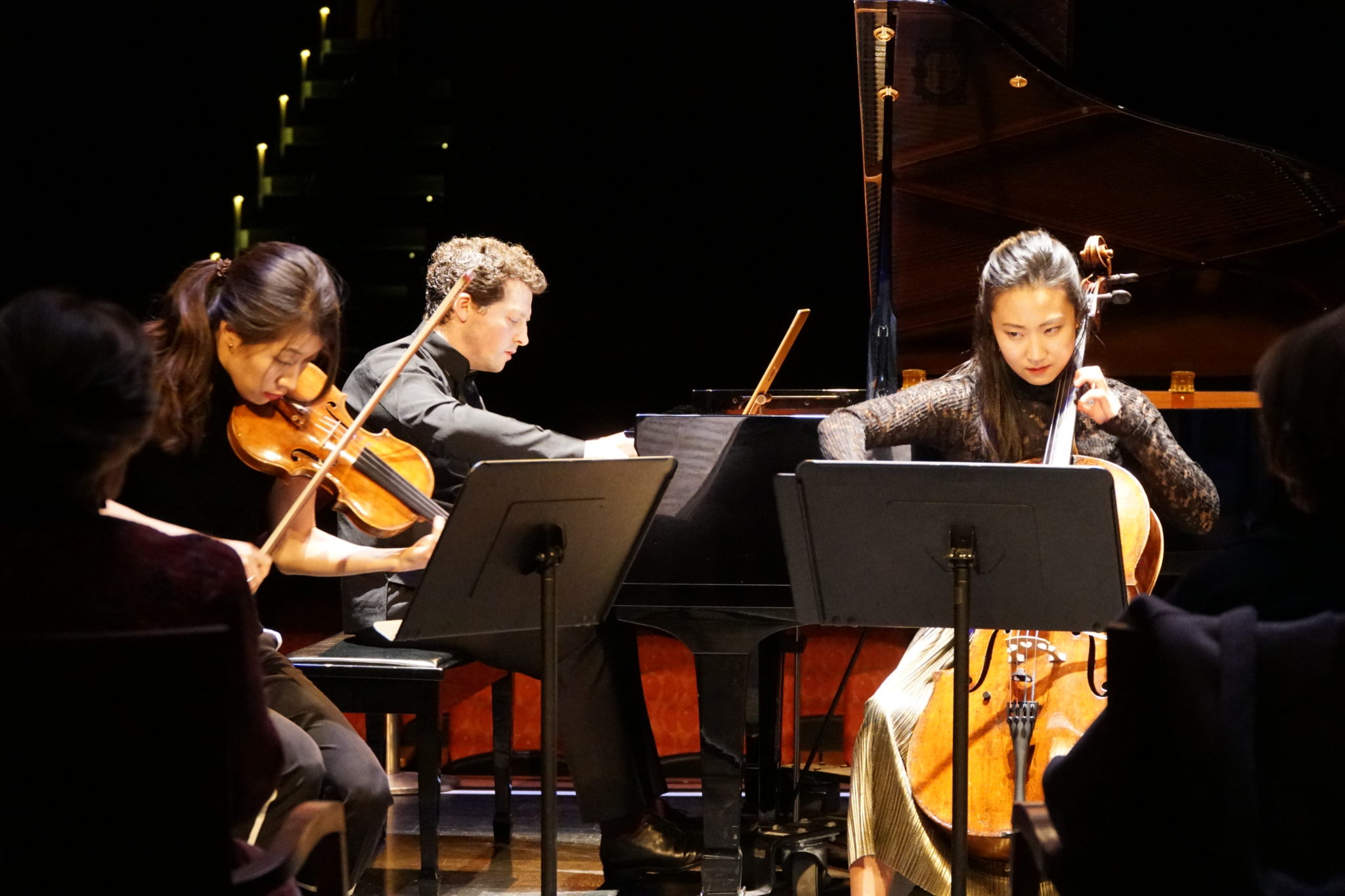 Performance photo with dark outline of audience menbers, Crystel Lee playing violin,  Henry Kramer playing piano, Jia Kim playing cello