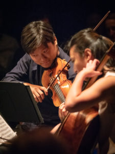 Spotlight on Siwoo Kim playing violin with Jia Kim playing cello, looking at music on stands with black background.