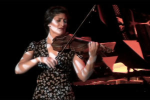 Michelle Ross playing violin in a black and white patterned dress onstage with specked lighting