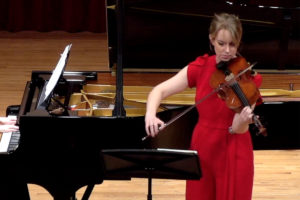 Molly Carr playing violin in a red dress standing in front of a piano.