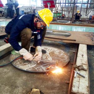 Harlan Mack on a residency in china wearing a dark blue coat and yello hard hat with safety glasses, crouching in a studio welding pieces together.