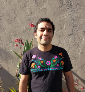 Gibran in a black t-shirt with pink and orange flowers, standing in front of a concrete wall with leafy plants with pink flowers behind him.