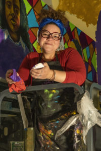 Photo of Jennifer Herrera Condry on a ladder with a mural behind her that she is working on - red shirt, dark rimmed glasses paint on her aprom.