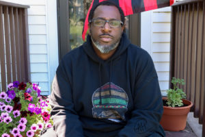 Photo of Will Kasso Condry wearing a hooded sweatshirt and glasses sitting on a porch with purple flowers to the left, a plant to the right and a red & black flag against a white house in the background.