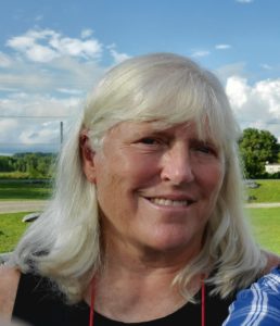 Photo of Belinda Emerson wearing a sleeveless black top, green lawn, blue skies and billowing clouds behind her