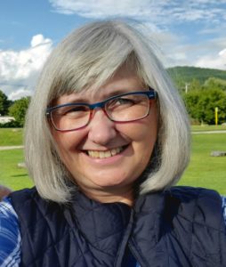 Photo of Julianne Nickerson wearing a blue flannel shirt and vest, green lawn, blue skies and billowing clouds behind her