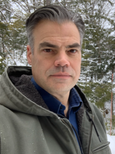Photo of Rich Miller in an olive hoodie outdoors in the winter with evergreens and snow