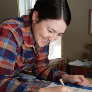 Amy Hook-Therrien wearing a multi-colored plaid shirt, brown hair in a braid smiling, working at a table drawing into a painting.