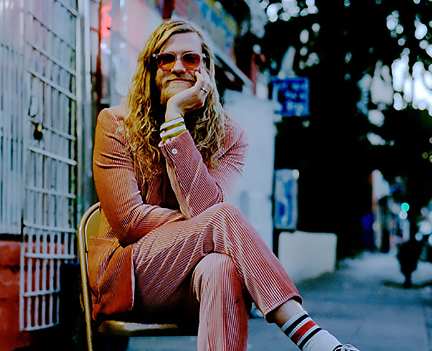Portrait of Allen Stone seated in a chair on the sidewalk in a pink suit with rose colored sunglasses.