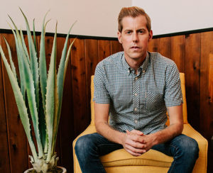 Portrait of Andrew McMahon sitting on yellow chair with tall green plant to his left