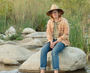 Portrait of musician Brett Dennen sitting riverside on a large rock wearing a hat - tall grasses behind him