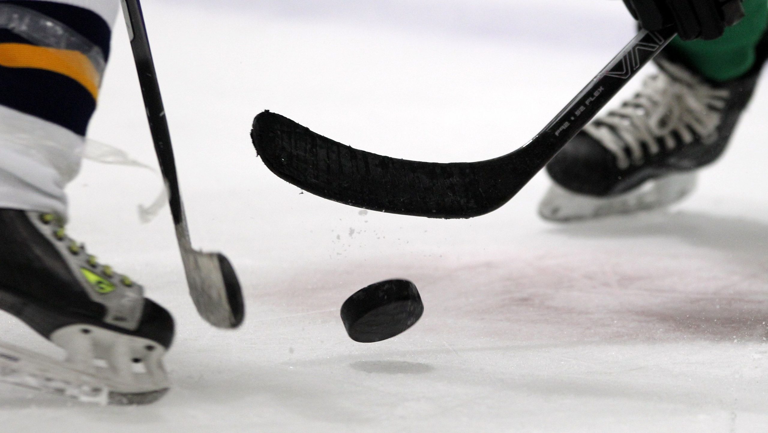 two hockey players, only skates visible, with two black hockey sticks going to hit a black hockey puck.