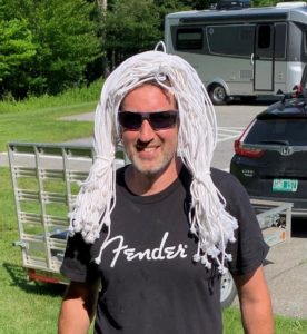 White man with black Fender t-shirt, black sunglasses, and white yarn mop on his head, in front of car trailer and back of car with VT license plate