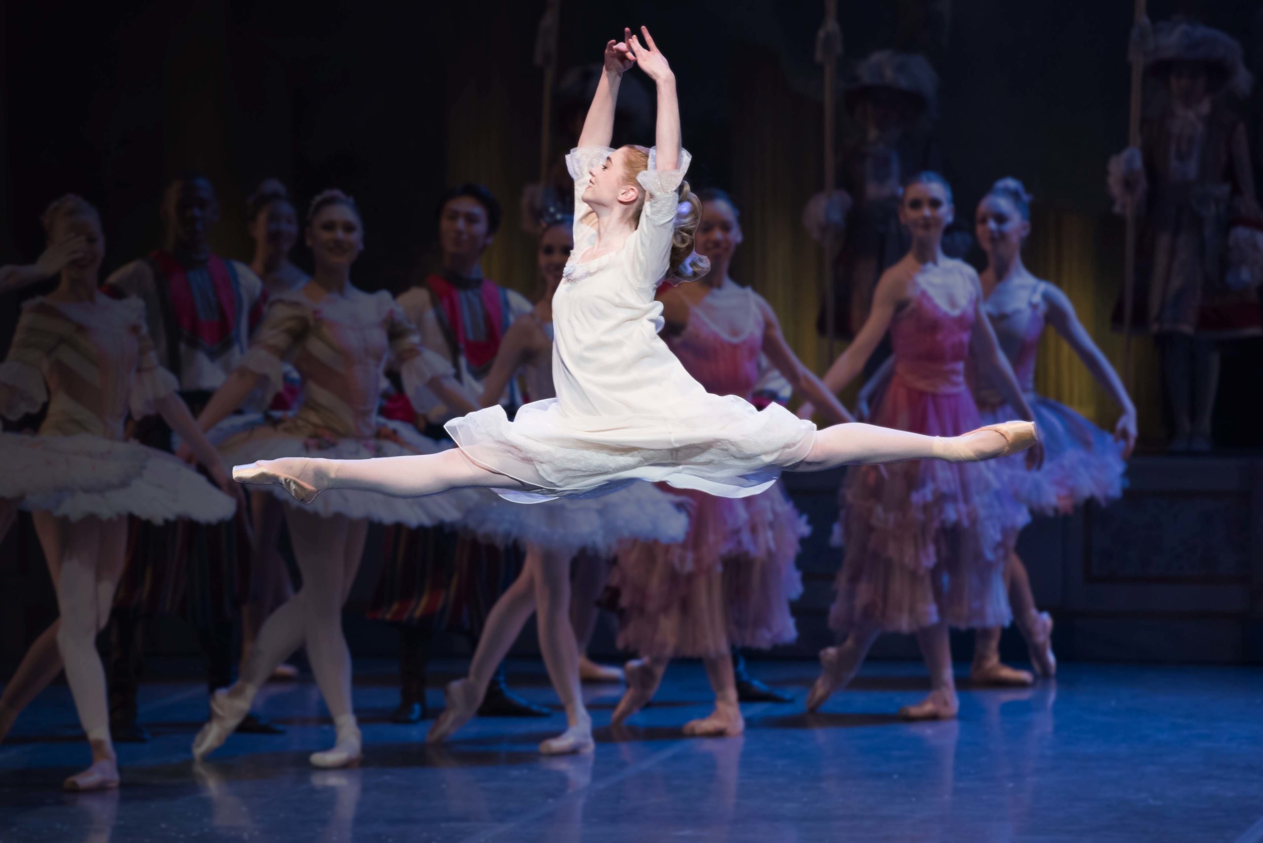 Ballerina leaping in full spit with arms held over her head, dressed in white nightgown, other dancers faded in the background