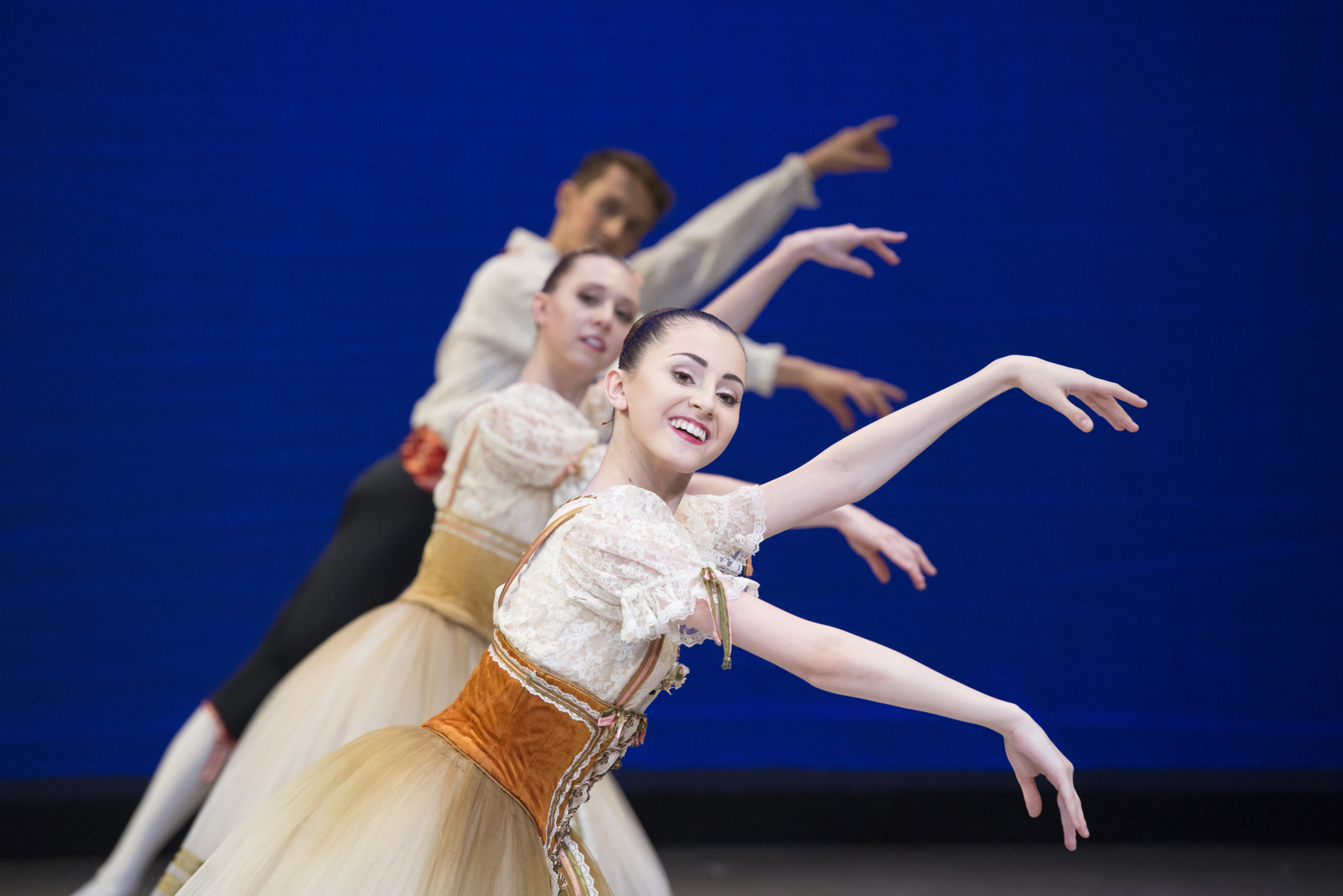 three ballet dancers in a line in front of each other with arms out to the right, hair in tights bun for the two females wearing beige tops and light organge tuille skirts and male in black pants and biege top