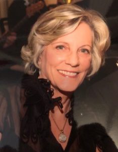 Headshot for Board Member Jill Witten, short coiffed blond hair, big smile, fancy black neckline blouse and necklace with large pendant jewel