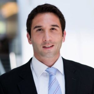 Headshot for Mirza Cavalic, dark hair, dark suit, white shirt, blue tie, blurry background