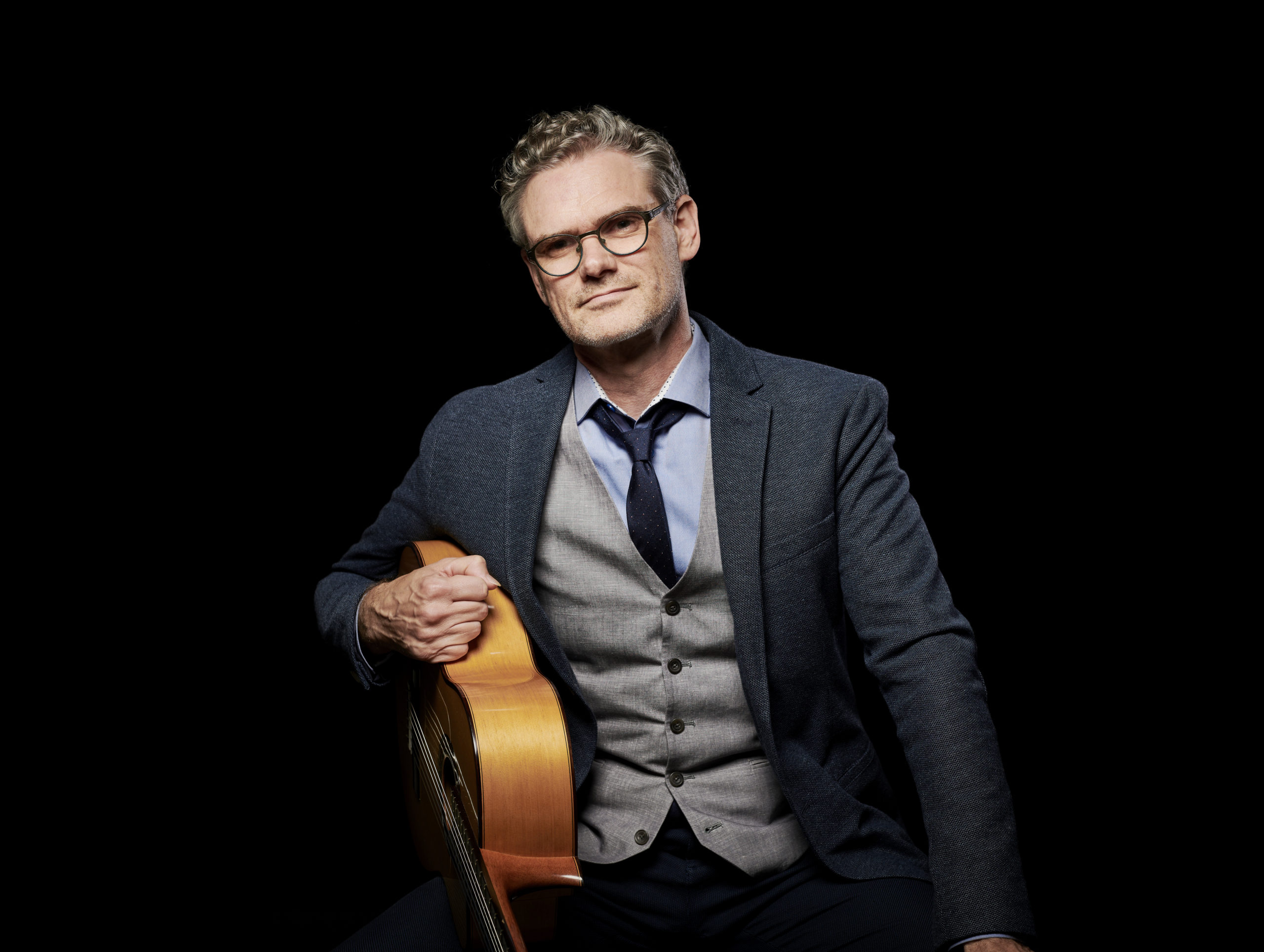 image of Jesse Cook, a man in a dark suit coat with black rimmed glasses holds a guitar under his right arm against a black background.