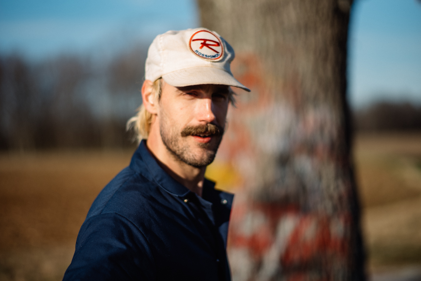 Rayland Baxter headshot - looking over hisright shoulder. Mustache, blonde hair sticking out of beige baseball cap