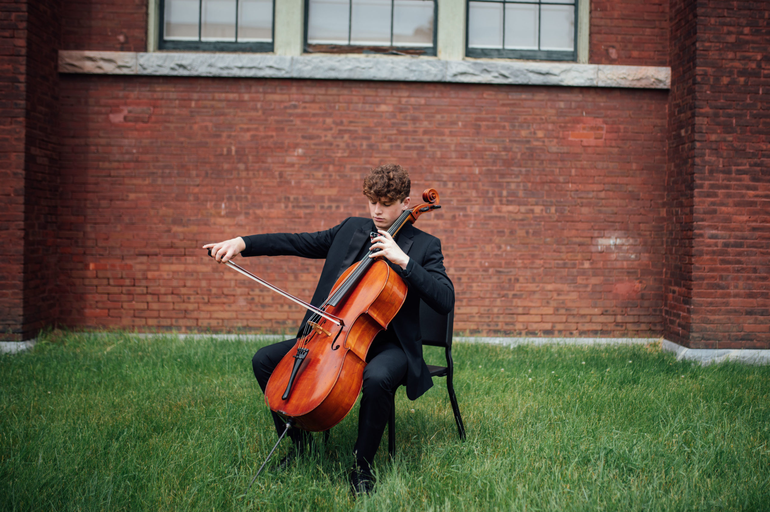 VYOA student musician is seated in front of a brick wall with cello, bow poised to play.