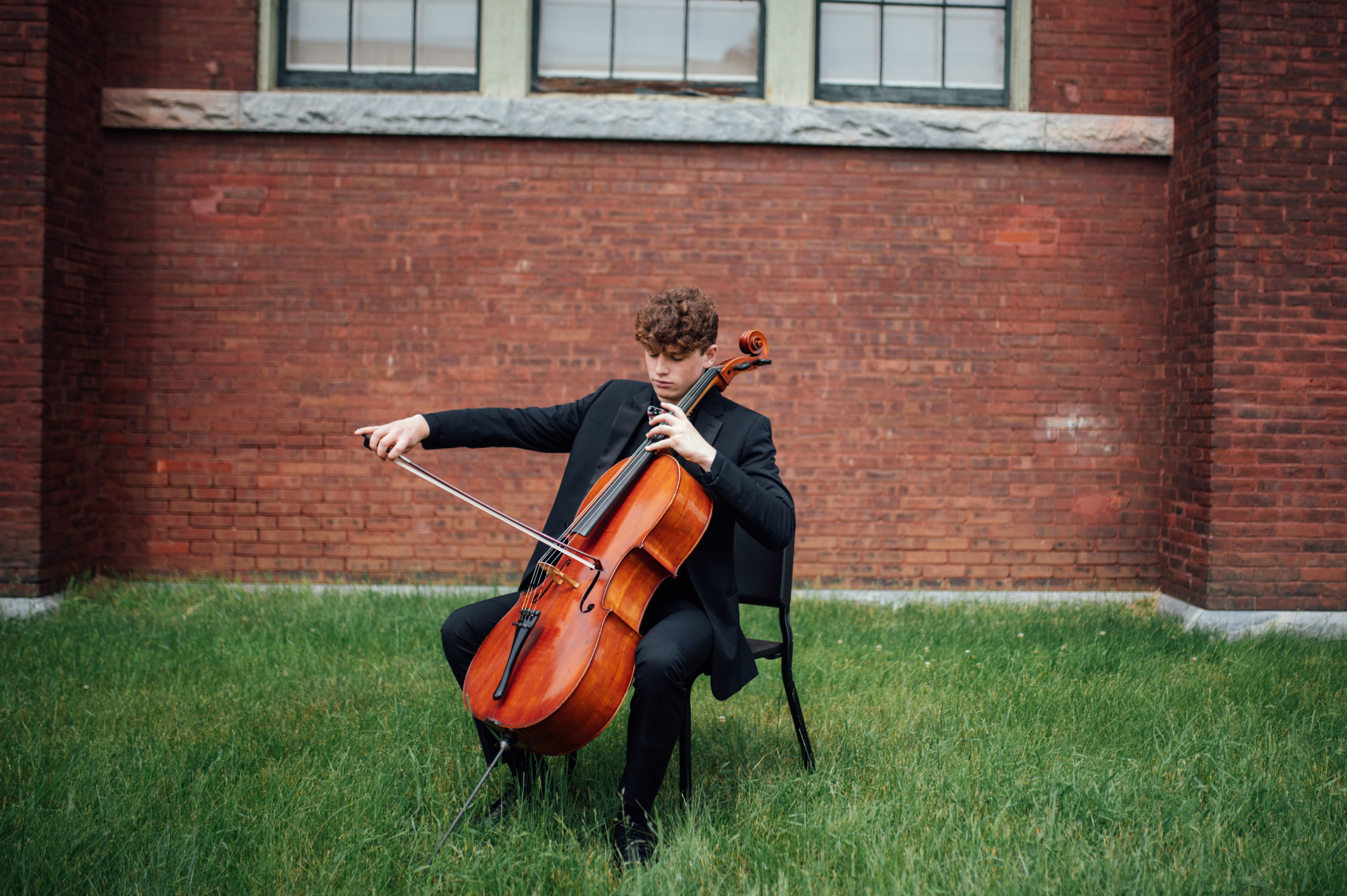 _DSC8702 VYOA student musician is seated in front of a brick wall with cello, bow poised to play.