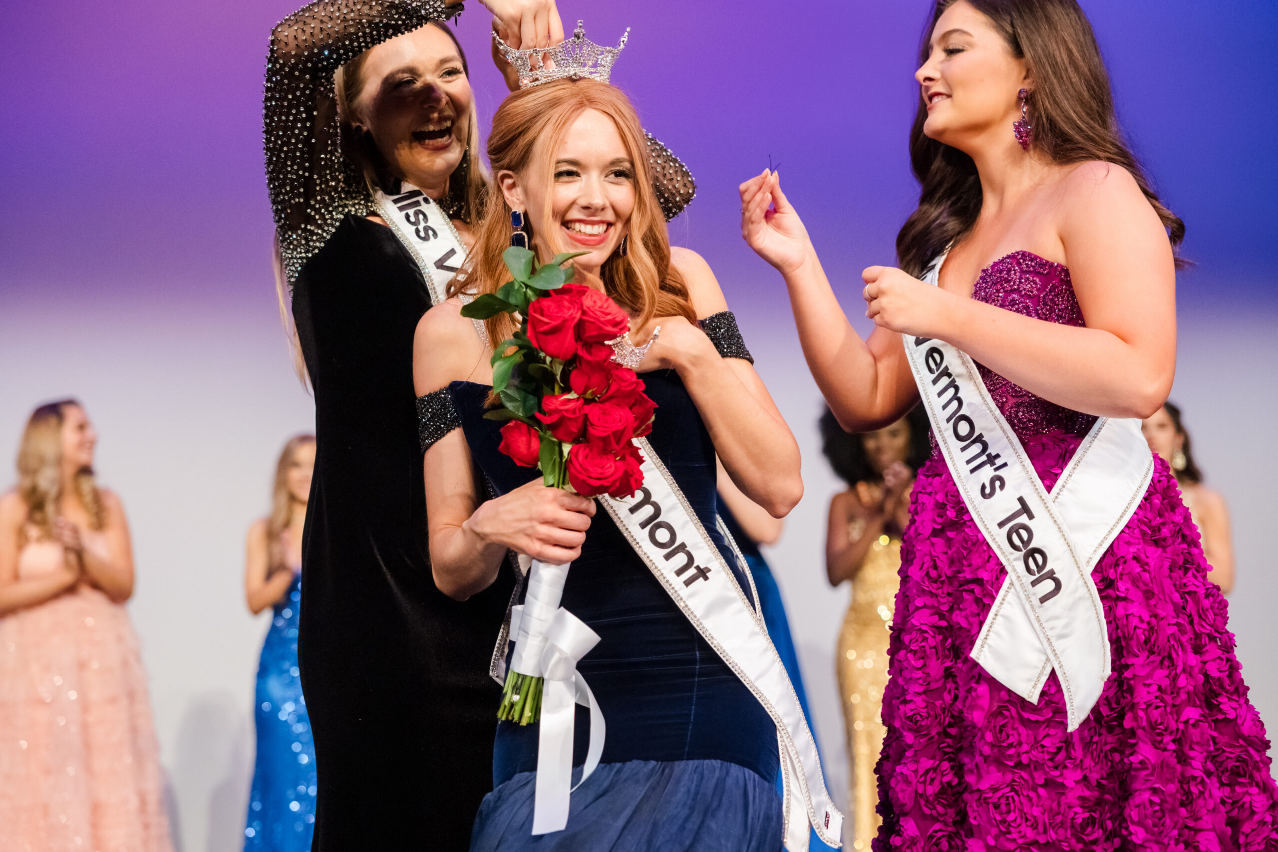 Women standing on stage in ball gowns. One in center is getting crowned